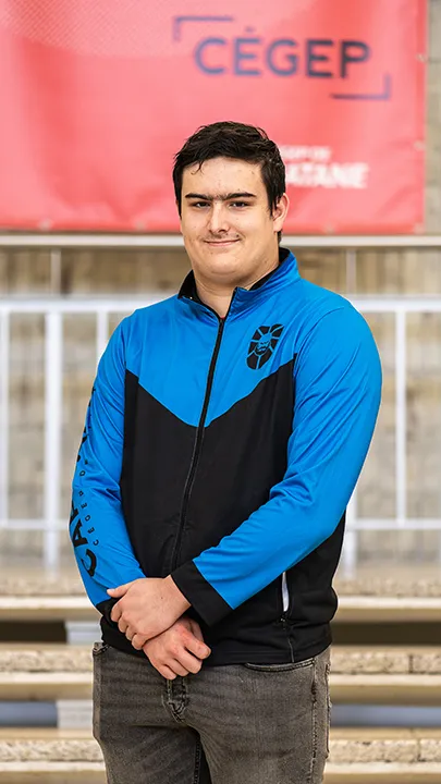 Un athlète de l'équipe de sport électronique pose sur les marches de l'escalier du hall d'entrée du Cégep de Matane pour sa photo d'équipe, vêtu de la veste bleue et noire des Capitaines.