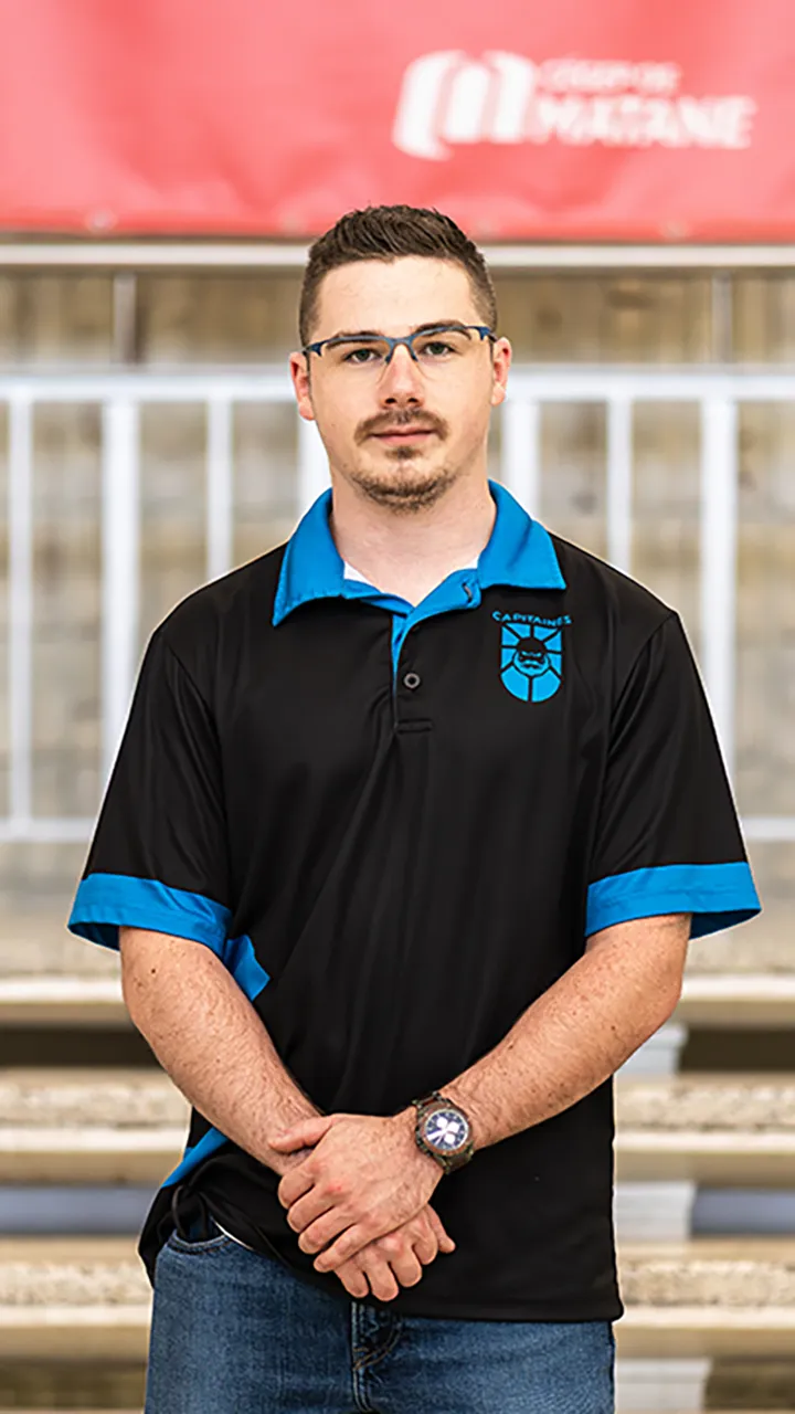 Un entraîneur vêtu du polo noir et bleu des Capitaines du Cégep de Matane pose dans les marches du puits du hall de l'établissement.