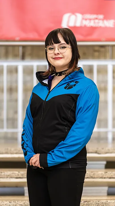 Une athlète de l'équipe de sport électronique pose sur les marches de l'escalier du hall d'entrée du Cégep de Matane pour sa photo d'équipe, vêtue de la veste bleue et noire des Capitaines.