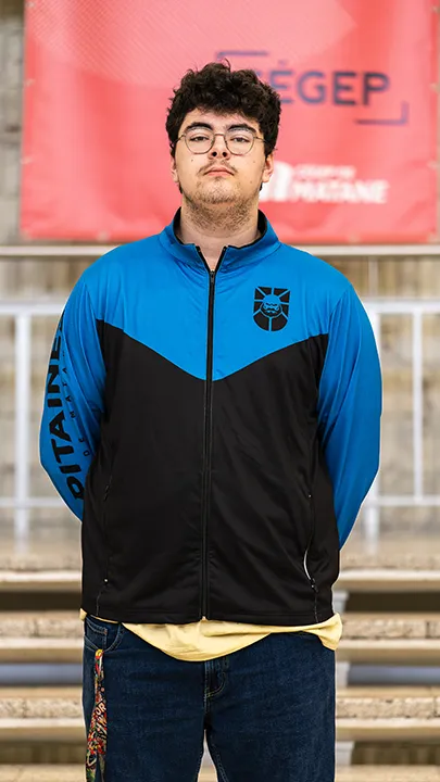 Un athlète de l'équipe de sport électronique pose sur les marches de l'escalier du hall d'entrée du Cégep de Matane pour sa photo d'équipe, vêtu de la veste bleue et noire des Capitaines.