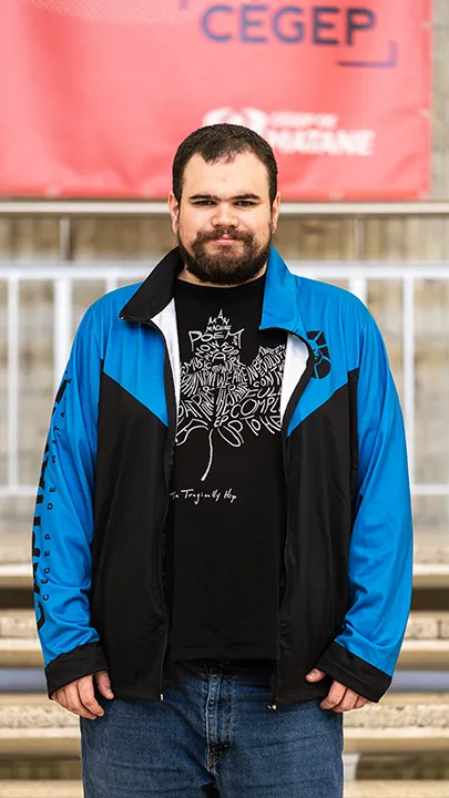 Un athlète de l'équipe de sport électronique pose sur les marches de l'escalier du hall d'entrée du Cégep de Matane pour sa photo d'équipe, vêtu de la veste bleue et noire des Capitaines.