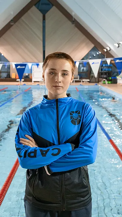 Une athlète des Capitaines, dans une piscine, sur le bord d'un bassin de natation, pose avec la tenue bleue et noire des Capitaines dans le cadre de la photo officielle de l'équipe collégiale de natation.