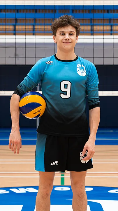 Dans un gymnase, devant un filet de volleyball installé sur un terrain omnisport, un athlète en tenue officielle des Capitaines pose avec un ballon de volleyball lors de la prise des photos officielles de l'équipe.