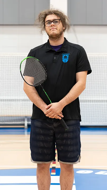 Sur un terrain de badminton, dans un gymnase, un athlète de l'équipe des Capitaines, en tenue officielle, une raquette de badminton à la main, prend la pose dans le cadre des photos de l'équipe.