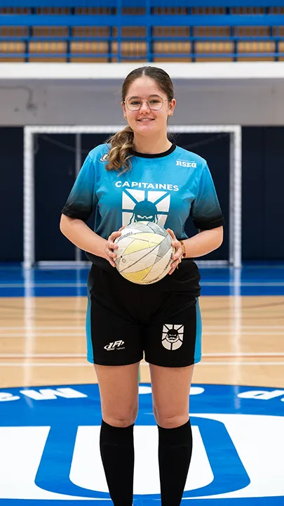 Dans un gymnase, une athlète étudiante du cégep pose seule, en tenue bleue et noire des Capitaines, un ballon à la main, dans le cadre des photos d'équipe de futsal féminin pour l'année 2024-2025.