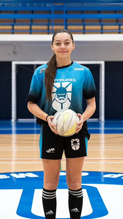 Dans un gymnase, une athlète étudiante du cégep pose seule, en tenue bleue et noire des Capitaines, un ballon à la main, dans le cadre des photos d'équipe de futsal féminin pour l'année 2024-2025.