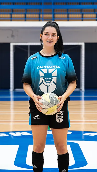 Dans un gymnase, une athlète étudiante du cégep pose seule, en tenue bleue et noire des Capitaines, un ballon à la main, dans le cadre des photos d'équipe de futsal féminin pour l'année 2024-2025.