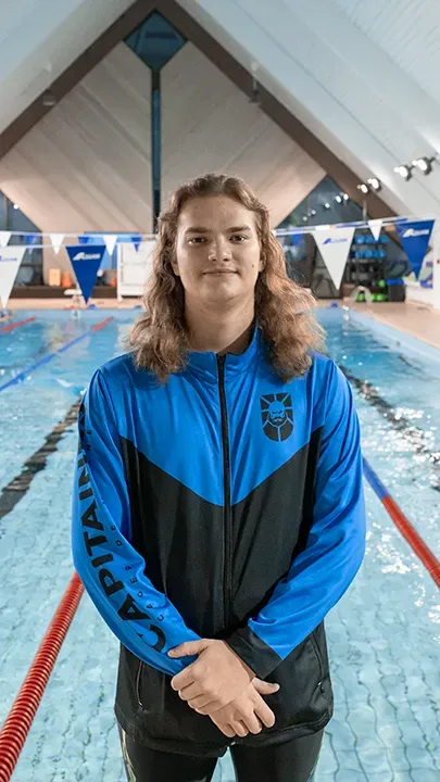 Un athlète des Capitaines, dans une piscine, sur le bord d'un bassin de natation, pose avec la tenue bleue et noire des Capitaines dans le cadre de la photo officielle de l'équipe collégiale de natation.