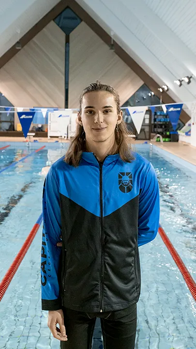 Un athlète des Capitaines, dans une piscine, sur le bord d'un bassin de natation, pose avec la tenue bleue et noire des Capitaines dans le cadre de la photo officielle de l'équipe collégiale de natation.