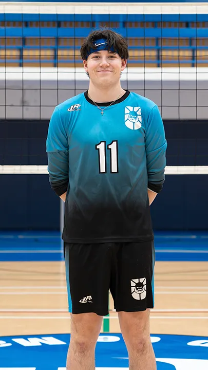 Dans un gymnase, devant un filet de volleyball installé sur un terrain omnisport, un athlète en tenue officielle des Capitaines pose lors de la prise des photos officielles de l'équipe.