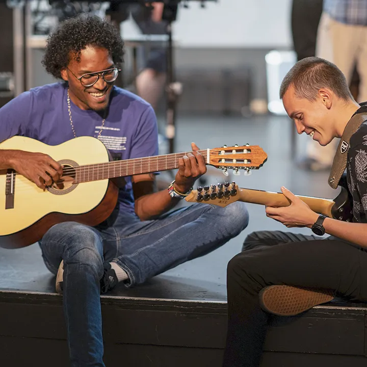 Deux étudiants assis l'un en face de l'autre, sur le bord d'une scène de salle de spectacle, jouent de la guitare ensemble dans le cadre du club des arts de la scène du cégep.