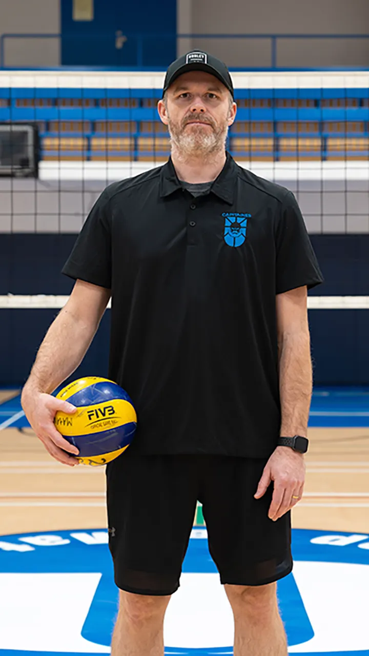 Sur le terrain omnisport d'un gymnase, devant un filet de volleyball, un homme en tenue d'entraîneur de volleyball prend la pose un ballon à la main dans le cadre des photos de l'équipe de volleyball masculin des Capitaines du cégep.