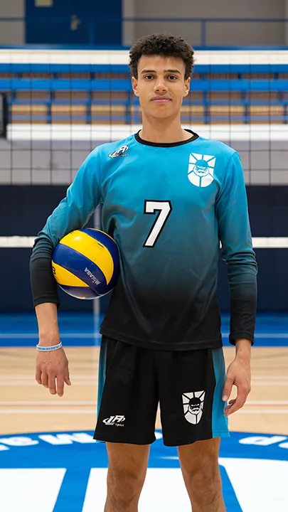 Dans un gymnase, devant un filet de volleyball installé sur un terrain omnisport, un athlète en tenue officielle des Capitaines pose avec un ballon de volleyball lors de la prise des photos officielles de l'équipe.