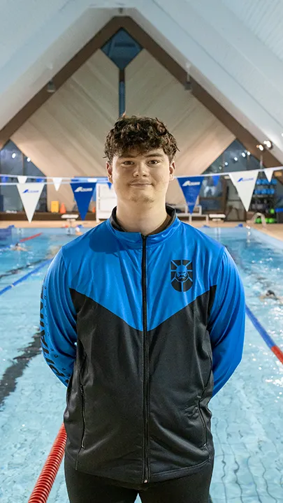 Un athlète des Capitaines, dans une piscine, sur le bord d'un bassin de natation, pose avec la tenue bleue et noire des Capitaines dans le cadre de la photo officielle de l'équipe collégiale de natation.