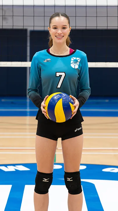 Sur le terrain omnisport d'un gymnase, devant un filet de volleyball, une étudiante vêtue de l'uniforme des Capitaines prend la pose, un ballon de volleyball entre les mains, dans le cadre des photos officielles de l'équipe féminine de volleyball du cégep.