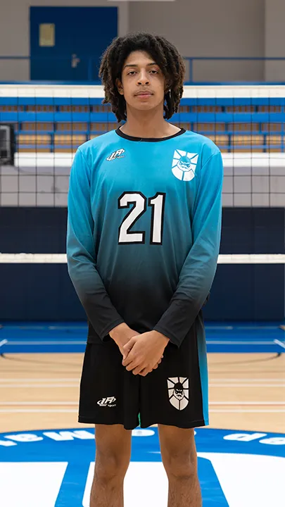 Dans un gymnase, devant un filet de volleyball installé sur un terrain omnisport, un athlète en tenue officielle des Capitaines pose lors de la prise des photos officielles de l'équipe.