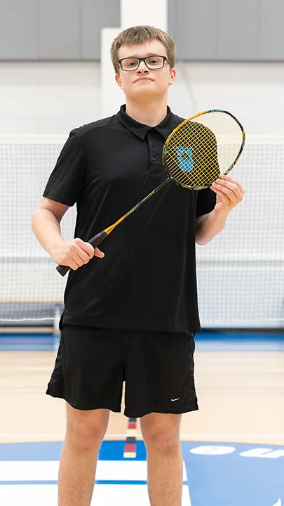 Sur un terrain de badminton, dans un gymnase, un athlète de l'équipe des Capitaines, en tenue officielle, une raquette de badminton à la main, prend la pose dans le cadre des photos de l'équipe.