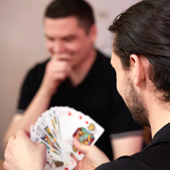 Une photo en gros plan montre deux personnes autour d'une table en train de jouer au tarot, jeu de carte français. Le jeu d'une des deux personnes est déployé dans sa main, tandis que son partenaire de jeu semble réfléchir à son prochain coup.