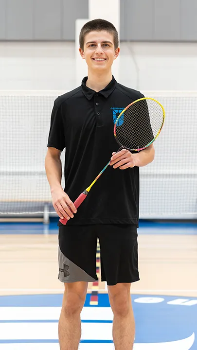 Sur un terrain de badminton, dans un gymnase, un athlète de l'équipe des Capitaines, en tenue officielle, une raquette de badminton à la main, prend la pose dans le cadre des photos de l'équipe.