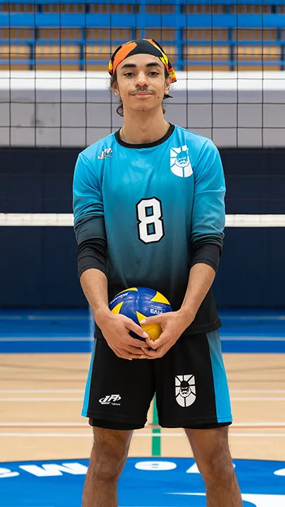 Dans un gymnase, devant un filet de volleyball installé sur un terrain omnisport, un athlète en tenue officielle des Capitaines pose avec un ballon de volleyball lors de la prise des photos officielles de l'équipe.