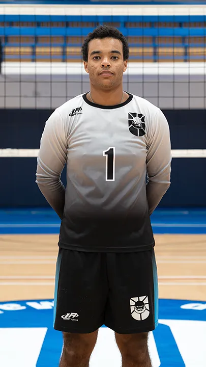 Dans un gymnase, devant un filet de volleyball installé sur un terrain omnisport, un athlète en tenue officielle des Capitaines pose lors de la prise des photos officielles de l'équipe.