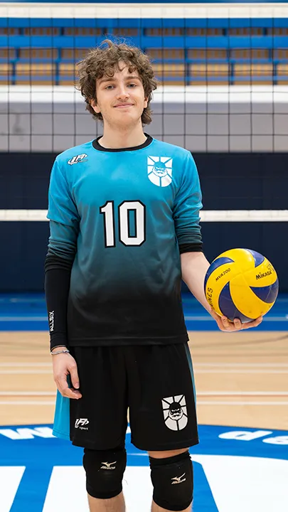 Dans un gymnase, devant un filet de volleyball installé sur un terrain omnisport, un athlète en tenue officielle des Capitaines pose avec un ballon de volleyball lors de la prise des photos officielles de l'équipe.