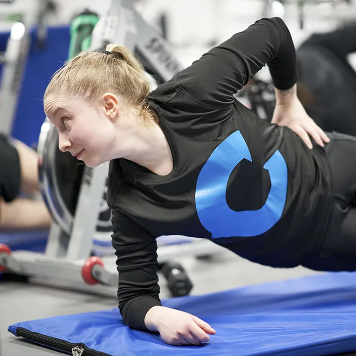 Une jeune femme, dans la vingtaine, effectue une planche latérale sur un tapis de sol dans le cadre d'un exercice de gainage musculaire lors d'une séance collective d'exercices au sol.