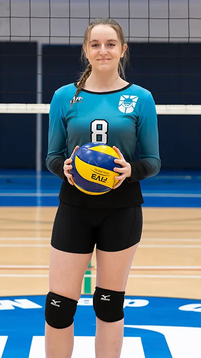 Sur le terrain omnisport d'un gymnase, devant un filet de volleyball, une étudiante vêtue de l'uniforme des Capitaines prend la pose, un ballon de volleyball entre les mains, dans le cadre des photos officielles de l'équipe féminine de volleyball du cégep.