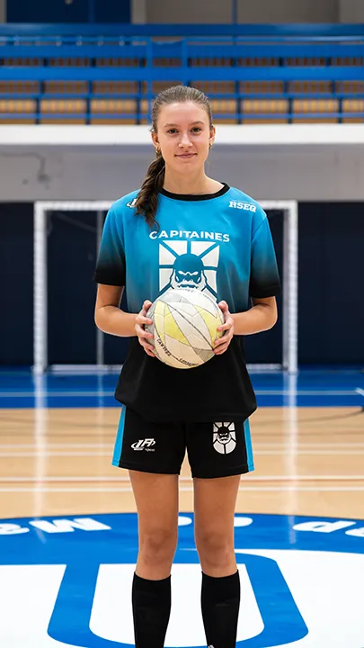 Dans un gymnase, une athlète étudiante du cégep pose seule, en tenue bleue et noire des Capitaines, un ballon à la main, dans le cadre des photos d'équipe de futsal féminin pour l'année 2024-2025.