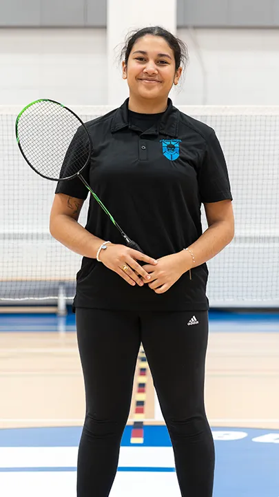 Sur un terrain de badminton, dans un gymnase, une athlète de l'équipe des Capitaines, en tenue officielle, une raquette de badminton à la main, prend la pose dans le cadre des photos de l'équipe.