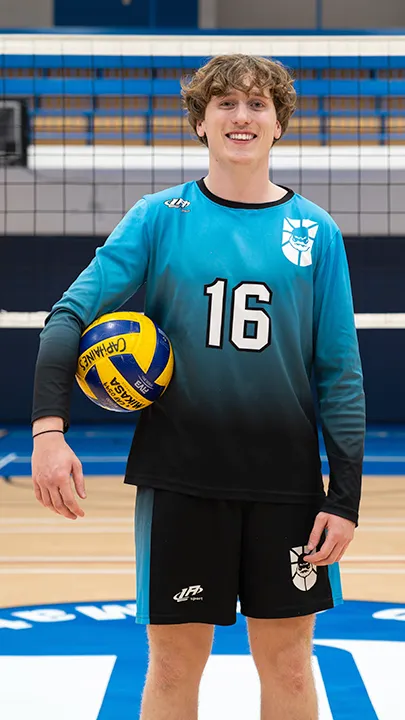 Dans un gymnase, devant un filet de volleyball installé sur un terrain omnisport, un athlète en tenue officielle des Capitaines pose avec un ballon de volleyball lors de la prise des photos officielles de l'équipe.