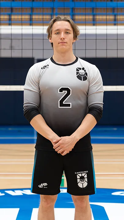 Dans un gymnase, devant un filet de volleyball installé sur un terrain omnisport, un athlète en tenue officielle des Capitaines pose lors de la prise des photos officielles de l'équipe.