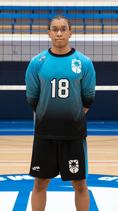 Dans un gymnase, devant un filet de volleyball installé sur un terrain omnisport, un athlète en tenue officielle des Capitaines pose lors de la prise des photos officielles de l'équipe.