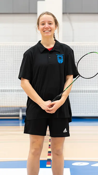 Sur un terrain de badminton, dans un gymnase, une athlète de l'équipe des Capitaines, en tenue officielle, une raquette de badminton à la main, prend la pose dans le cadre des photos de l'équipe.