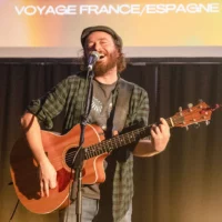 Un guitariste barbu est en train de chanter, en chemise à carreaux et une casquette en laine sur la tête.